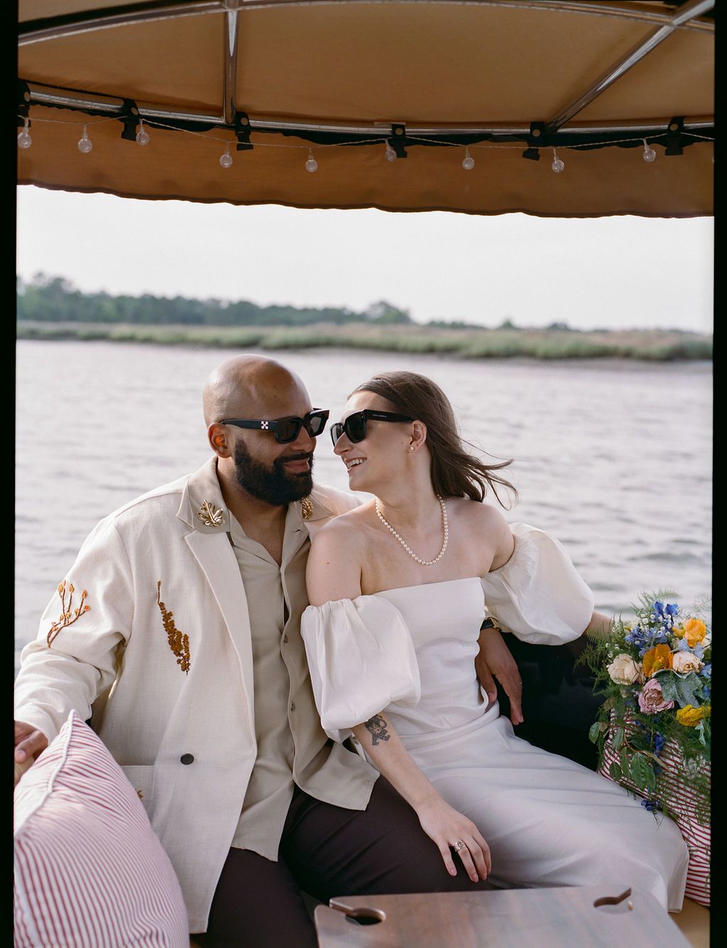 Newlyweds leaning in close under the Joyride canopy, bouquet beside them, marsh behind