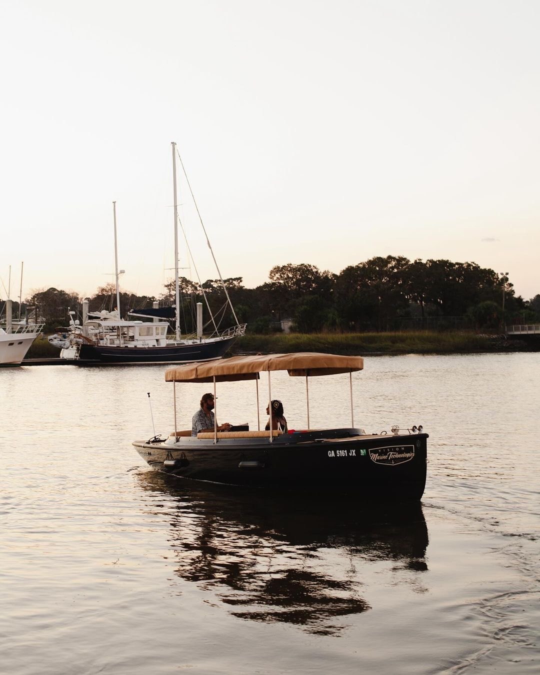 The Joyride electric boat gliding across calm water at sunset, two guests under the tan canopy, sailboats and marsh behind